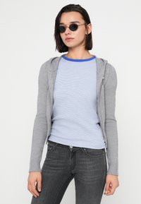 Gray zip-up hoodie with long sleeves over a blue and white striped T-shirt. Model wears black round sunglasses and dark gray jeans.