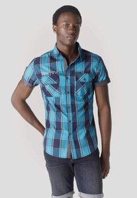 Młody mężczyzna o krótkich włosach, ubrany w niebieską kratowaną koszulę z krótkim rękawem i ciemne, podwinięte dżinsy, stoi z jedną ręką za plecami.