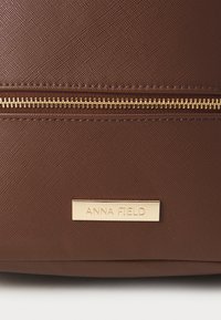 Sac à main brun texturé avec une fermeture éclair dorée et une plaque rectangulaire portant le nom "ANNA FIELD". Design robuste avec des détails de couture visibles.