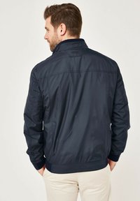 Navy blue bomber jacket with a smooth, shiny texture, ribbed cuffs, and collar, features logo stitching on the back and side pockets.