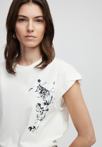 Witte katoenen t-shirt met korte opgerolde mouwen. Heeft een zwart-wit bloemenafdruk op de linkerschouder.