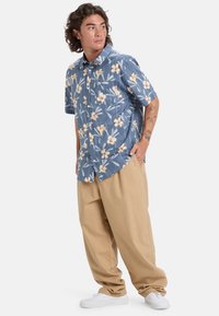 Košile s krátkým rukávem modré barvy s bílými a žlutými květinami, doplněná volnými kalhotami ve světle béžové barvě a bílými teniskami.