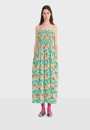 Woman with long dark hair facing forward wearing a sleeveless, smocked, ankle-length dress with a colorful floral and leaf pattern.