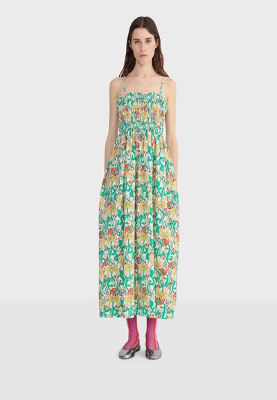 Femme aux longs cheveux foncés regardant de face, portant une robe sans manches, smockée, longueur cheville, avec un motif coloré de fleurs et de feuilles.