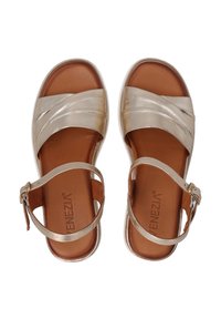 Beige sandalen met gekruiste, metallic banden en een achterste enkelband. Heeft een bruine binnenzool en een licht verhoogde witte zool.