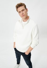 Man in een witte half-zip sweatshirt, donkere broek en witte sneakers, staand met de handen in de zakken tegen een effen achtergrond.