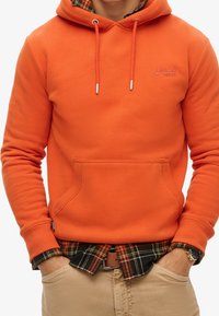 Sudadera con capucha naranja con bolsillo frontal, cordones contrastantes y logo. Llevada sobre una camisa de franela a cuadros, combinada con pantalones caqui.