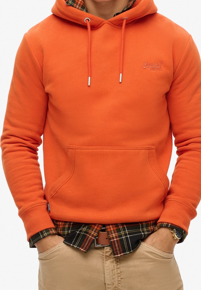 Sudadera con capucha naranja con bolsillo frontal, cordones contrastantes y logo. Llevada sobre una camisa de franela a cuadros, combinada con pantalones caqui.