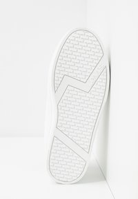 Semelle de basket blanche présentant un motif de grip texturé, un détail de logo et un design à bord arrondi, affichée sur un fond clair.