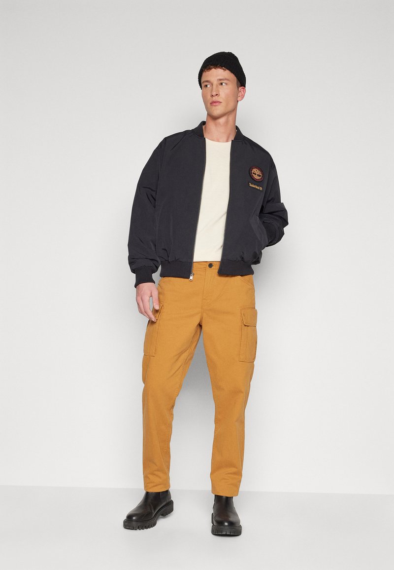 Czarna kurtka bomberka z prążkowanymi mankietami, beżowa koszula pod spodem oraz ochrowe spodnie cargo z bocznymi kieszeniami; model w czarnych botkach Chelsea.