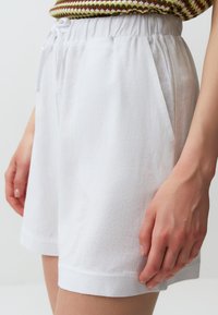 Pantaloni corti bianchi con vita elastica e cordoncino, dotati di tasche laterali e orlo ripiegato. Il tessuto ha una finitura testurizzata.