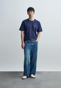 Μπλε ναυτικό μπλουζάκι με ελαφρύ γραφικό σχέδιο, συνδυασμένο με ανοιχτό μπλε τζιν παντελόνι φαρδιάς γραμμής και λευκά αθλητικά παπούτσια. Λείο υλικό, casual εφαρμογή.