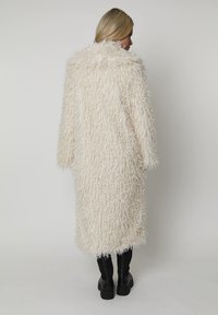 Długi, puszysty płaszcz w kolorze kremowym z teksturowanej, kręconej tkaniny. Posiada opadające ramiona i szeroki kołnierz, zestawiony z wysokimi czarnymi botkami.