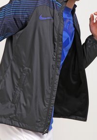 Chaqueta negra ligera con cierre de cremallera, con rayas horizontales azules en los hombros y bolsillos laterales. El forro interior es oscuro.