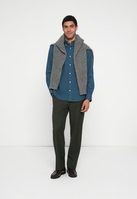 Sivá pletená vesta cez modrú košeľu s gombíkmi, doplnená o olivovo zelené nohavice a tmavohnedé topánky, na jednoduchom pozadí.