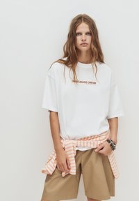 Weißes T-Shirt mit dem Text "ICH BRAUCHE EINE EISCREME" in Braun, getragen mit khakifarbenen Shorts. Gestreifter orangener Stoff um die Taille gebunden.