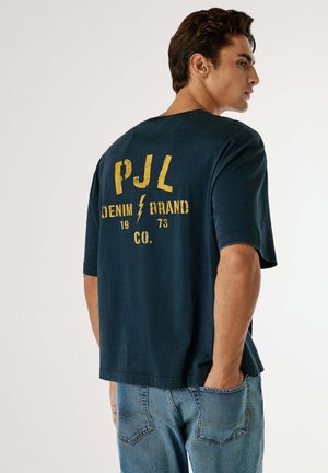 Navy blauwe oversized T-shirt met gele tekst "PJL DENIM BRAND CO. 1973" en een bliksemschichtontwerp, gemaakt van zachte katoen.