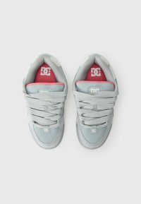 Ett par ljusgrå DC-märkes sneakers med vita och röda sulor, vända uppåt på en enkel vit bakgrund.
