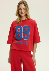 Młoda kobieta w czerwonej, luźnej koszuli z dużym niebieskim numerem 89 i napisem BLOOVII, w czerwonych spodniach do kompletu, uśmiechająca się do aparatu.