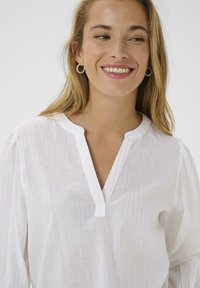 Weiße Bluse mit dezenten vertikalen Streifen, V-Ausschnitt und Knopfdetails. Weicher Stoff mit leichter, luftiger Textur und Ballonärmeln.
