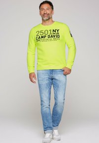 Sudadera de manga larga amarilla neón con texto gráfico en negro; combinada con jeans de mezclilla azul claro y zapatillas blancas. Diseño simple y casual.