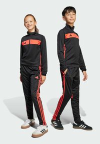 Svarta sportiga träningsdräkter med orangea ränder, zip-up jackor och byxor med muddar. Modellerna bär olika skor: en svart med vita accenter, en vit.