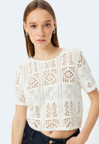 Top corto de punto de ganchillo blanco con mangas cortas, que presenta patrones abiertos, bordes de volantes y una forma cuadrada. Se combina con denim oscuro.