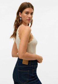 Top senza maniche lavorato a maglia color crema con scollatura a zig zag e motivo a intreccio aperto, abbinato a jeans scuri a vita alta con etichetta in pelle.