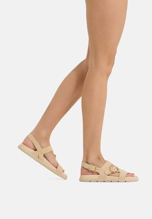 Beine mit beigen flachen Sandalen mit zwei Schnallenriemen, dargestellt beim Gehen vor einem einfarbigen hellen Hintergrund.