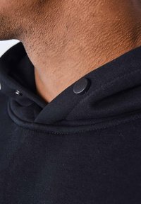 Cuello de sudadera negra con un tejido suave y textura notable, acentuado por un botón a presión negro y un diseño de capucha en capas.