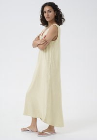 Robe maxi côtelée, jaune clair, avec une coupe décontractée, des fentes latérales et de larges emmanchures. Associée à des tongs blanches et des accessoires dorés.