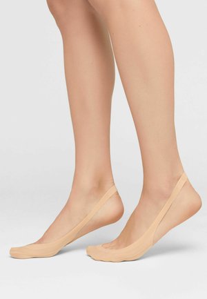 Par de piernas desnudas con calcetines invisibles color nude con correas en el talón sobre un fondo claro y liso.