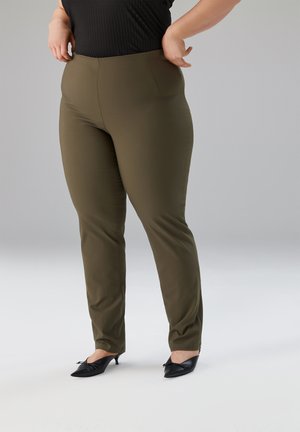 Frau trägt olivgrüne eng anliegende Hose und schwarze Schuhe mit kleinem Absatz, steht mit den Händen in der Hüfte vor einfarbigem Hintergrund.