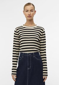 Haut à manches longues rayé en noir et crème, en tissu tricoté avec une texture côtelée. Associé à une jupe en denim foncé avec des coutures blanches.