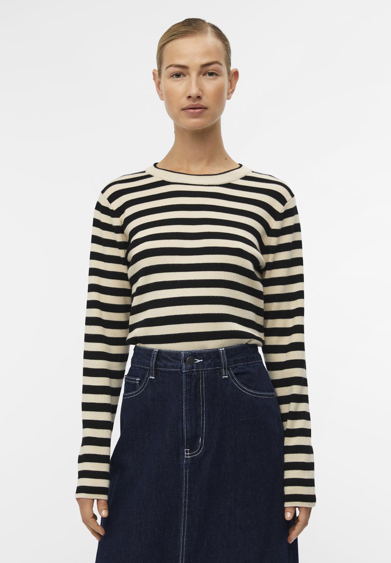 Haut à manches longues rayé en noir et crème, en tissu tricoté avec une texture côtelée. Associé à une jupe en denim foncé avec des coutures blanches.