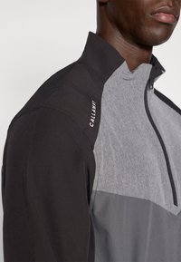 Halv-zip jakke i svart, grått og lyse grått materiale med en teksturert øverste seksjon; har en Callaway-logo på skulderen.