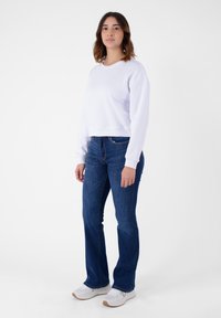 Felpa bianca corta con maniche lunghe, abbinata a jeans svasati blu scuro e sneakers grigie chiare su uno sfondo bianco semplice.