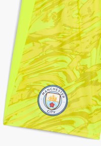 Camicia gialla brillante con un motivo testurizzato e ondulato. Presenta un logo circolare del Manchester City in basso a destra con accenti blu e rossi.