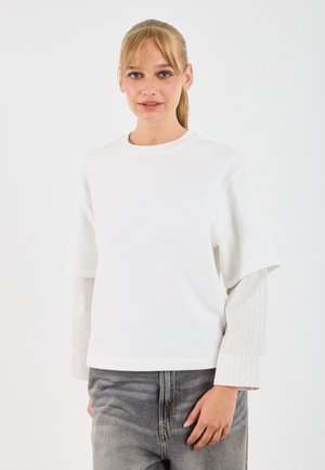 White short-sleeve top with a round neckline, layered over a light gray striped long-sleeve shirt. Model wears gray denim, hands relaxed.