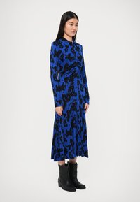 Hosszú ujjú ruha kék és fekete állatmintával, redőzött elülső részlettel és légies, áramló sziluettel, fekete bokacsizmával párosítva.