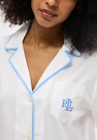 Γυναίκα με σγουρά μαλλιά που φοράει ένα λευκό πουκάμισο με κουμπιά και ανοιχτό μπλε διακόσμηση, με κεντημένο λογότυπο «RL» στην τσέπη στο στήθος.