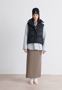 Svart puffervest over en lys blå skjorte, med en beige blyantskjørt og hvite sneakers. Outfiten er fullført med en svart lue.