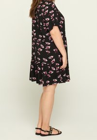 Kvinna som bär en svart blommig knälång klänning och svarta flat sandals med nitar, stående mot en ljus bakgrund.