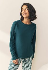 Türkises Langarmshirt mit Rundhalsausschnitt und Raglanärmeln, kombiniert mit grünen Leggings mit einem aufwendigen weißen Muster.