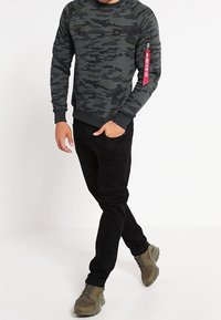 Felpa grigia con motivo camouflage a maniche lunghe, pantaloni neri e sneakers verdi. Presenta una tasca sul braccio con un'etichetta rossa.