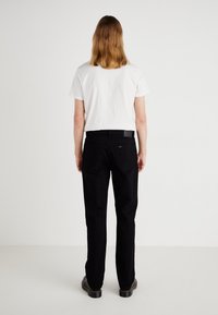 Õlgpikkade juustega inimene, kellel on seljas valge T-särk, mustad teksad ja mustad saapad, seisab seljaga sileda valge seina poole.