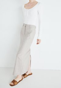 Person trägt ein weißes langärmeliges Oberteil, einen beige hochtaillierten Rock mit seitlichem Schlitz und braune Slip-On-Sandalen vor weißem Hintergrund.