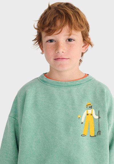 Jeune enfant roux avec des taches de rousseur portant un sweat-shirt vert avec un dessin de fermier en salopette jaune tenant une fourche.