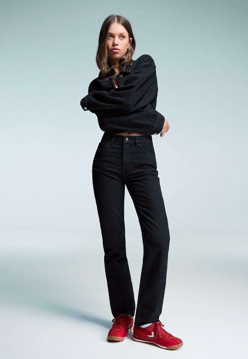 Črna denimska jakna in visoko pasne črne kavbojke, v kombinaciji z rdečimi supergami. Gladka tekstura, klasičen kroj in minimalističen dizajn.