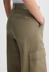 Olivgrüne Cargo-Hosen mit glatter Textur, seitlichen Taschen, einem nahtlosen Bund und einem kleinen Metallschriftzug.